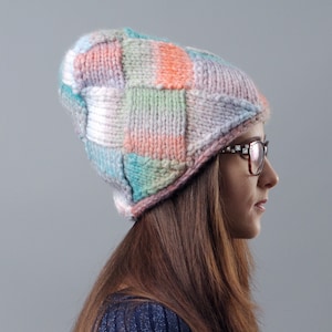 Può includere: Un berretto lavorato a maglia con un design patchwork nei toni pastello, tra cui rosa, blu, arancione e verde. Il berretto è indossato da una persona con lunghi capelli castani e occhiali. Il berretto sembra fatto a mano.