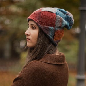 May include: A woman wearing a brown sweater and a colorful, patchwork knit beanie. The beanie features squares of red, blue, and other colors. The background is blurred, suggesting an outdoor setting with trees.