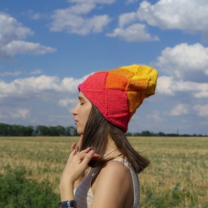 May include: A knitted beanie featuring a gradient pattern of diamond-shaped segments in vibrant shades of pink, orange, and yellow. The hat is constructed with a textured, geometric stitch pattern that transitions smoothly between the warm colour tones.