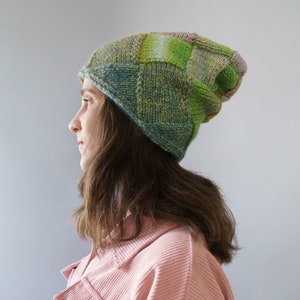 Könnte beinhalten: Handgestrickte Beanie-Mütze mit Patchwork-Design. Sie hat Quadrate aus grüner, blauer, gelber und violetter Wolle. Die Person, die sie trägt, hat braune Haare und trägt eine rosa Cordjacke.