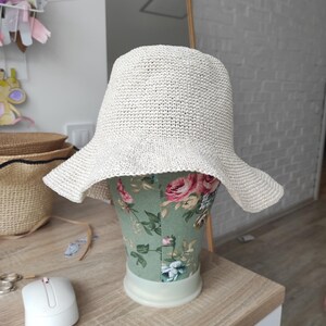 May include: A cream-colored, woven bucket hat with a wide, slightly flared brim. The hat features a textured, straw-like material and is displayed on a mannequin head covered in a green fabric with a pink and red floral pattern.