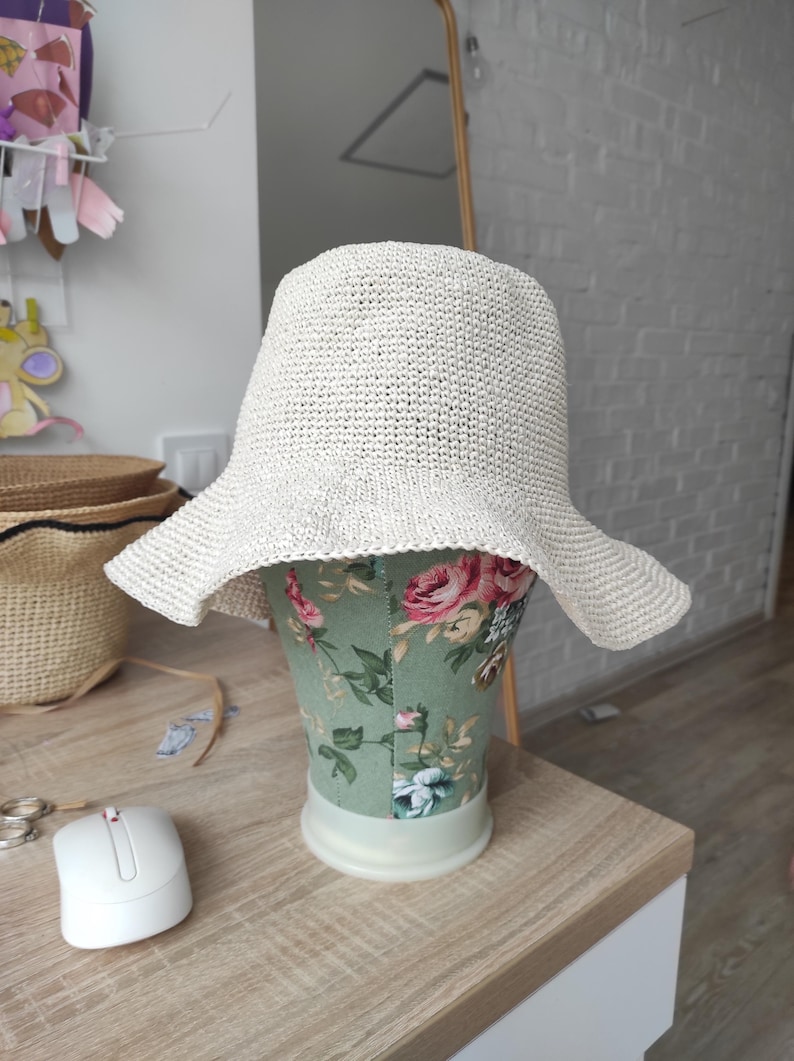 May include: A cream-colored, woven bucket hat with a wide, slightly flared brim. The hat features a textured, straw-like material and is displayed on a mannequin head covered in a green fabric with a pink and red floral pattern.