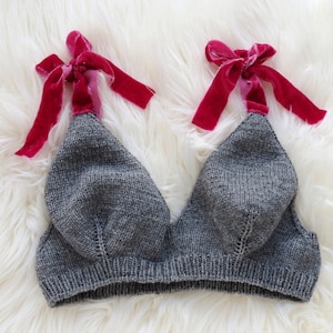 May include: A gray knitted bralette with red velvet ribbon ties. The bralette is made of a soft, textured knit fabric. The red ribbon adds a pop of color and a touch of elegance to the design. The bralette is displayed on a white, fluffy surface.