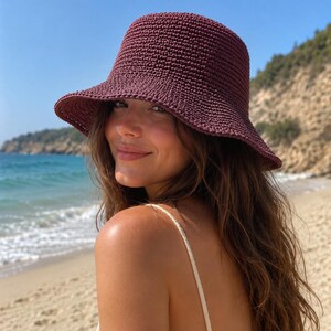 May include: A dark maroon bucket hat featuring a dense, textured crochet knit construction with a rounded crown and a slightly downturned brim. The hat is crafted from a solid-colored yarn, showcasing a consistent, tight stitch pattern throughout.