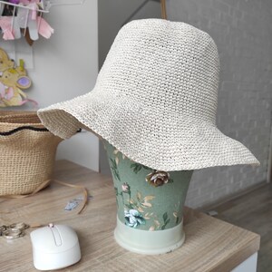 May include: A cream-colored bucket hat with a wide, slightly wavy brim, crafted from a textured, woven material. The hat is displayed on a green mannequin head featuring a floral pattern with pink and white roses and green leaves.