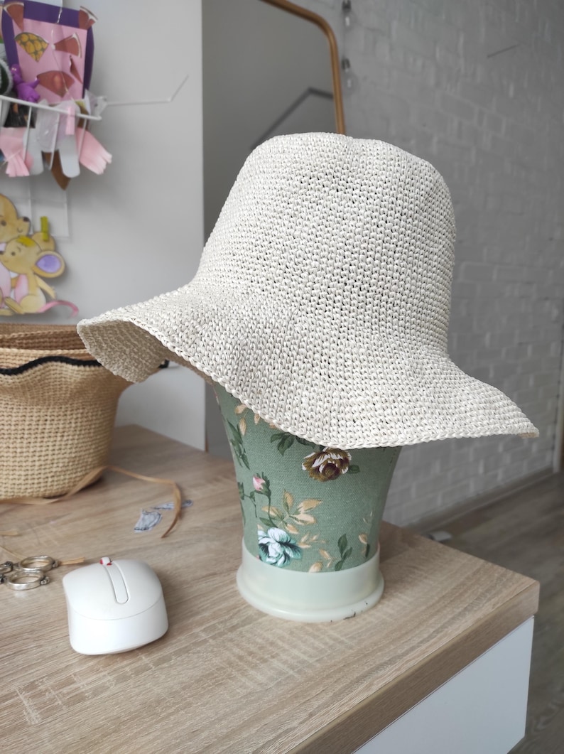 May include: A cream-colored bucket hat with a wide, slightly wavy brim, crafted from a textured, woven material. The hat is displayed on a green mannequin head featuring a floral pattern with pink and white roses and green leaves.