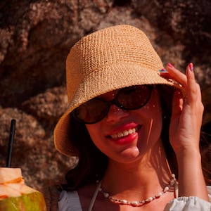 May include: A woman wearing a straw bucket hat and sunglasses smiles while holding the brim of the hat. She is wearing a white top and a necklace. A coconut drink with a black straw is in the foreground.