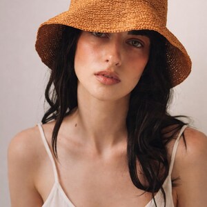 May include: A textured, rust-colored crochet bucket hat with a rounded crown and a downward-sloping brim. The hat features a dense, woven pattern throughout, providing a structured yet flexible shape that sits comfortably on the head.