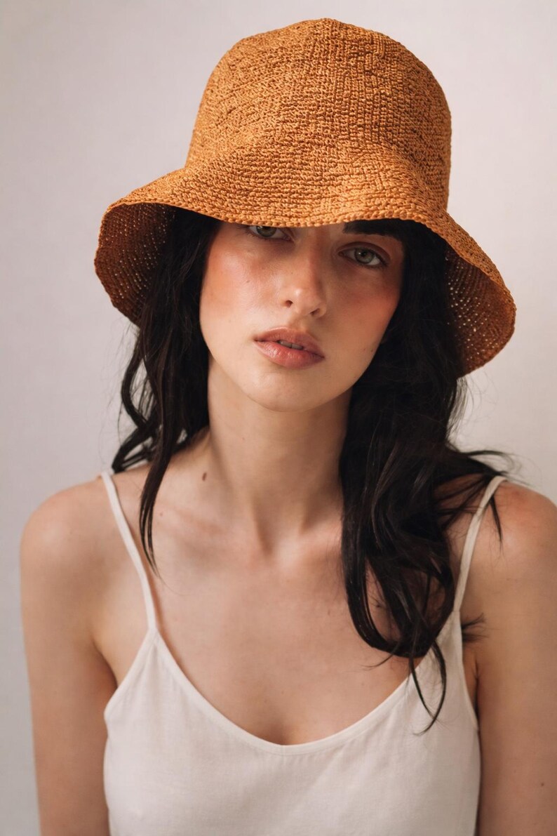 May include: A textured, rust-colored crochet bucket hat with a rounded crown and a downward-sloping brim. The hat features a dense, woven pattern throughout, providing a structured yet flexible shape that sits comfortably on the head.