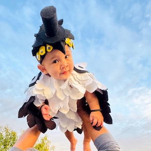 May include: A baby wearing a handmade bird costume with a black and white feathered body and a matching headpiece. The headpiece has yellow accents and a black top. The baby is being held up against a blue sky.