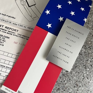 Puede incluir: Una tarjeta plateada con el texto "The US Olympic Team Watch. The Only Swiss Allowed On The 1996 US Olympic Team." La tarjeta está sobre una bandera estadounidense roja, blanca y azul.