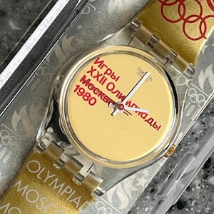 Puede incluir: Reloj Swatch vintage con correa dorada con los anillos olímpicos y la palabra "Olympia". La esfera es amarilla con texto ruso rojo. El reloj está en su embalaje original.