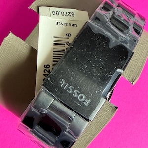 May include: A Fossil brand watch with a silver-tone metal band and a black watch face, still in its original packaging. The watch is partially covered in plastic. The watch is in a tan box with a price tag.