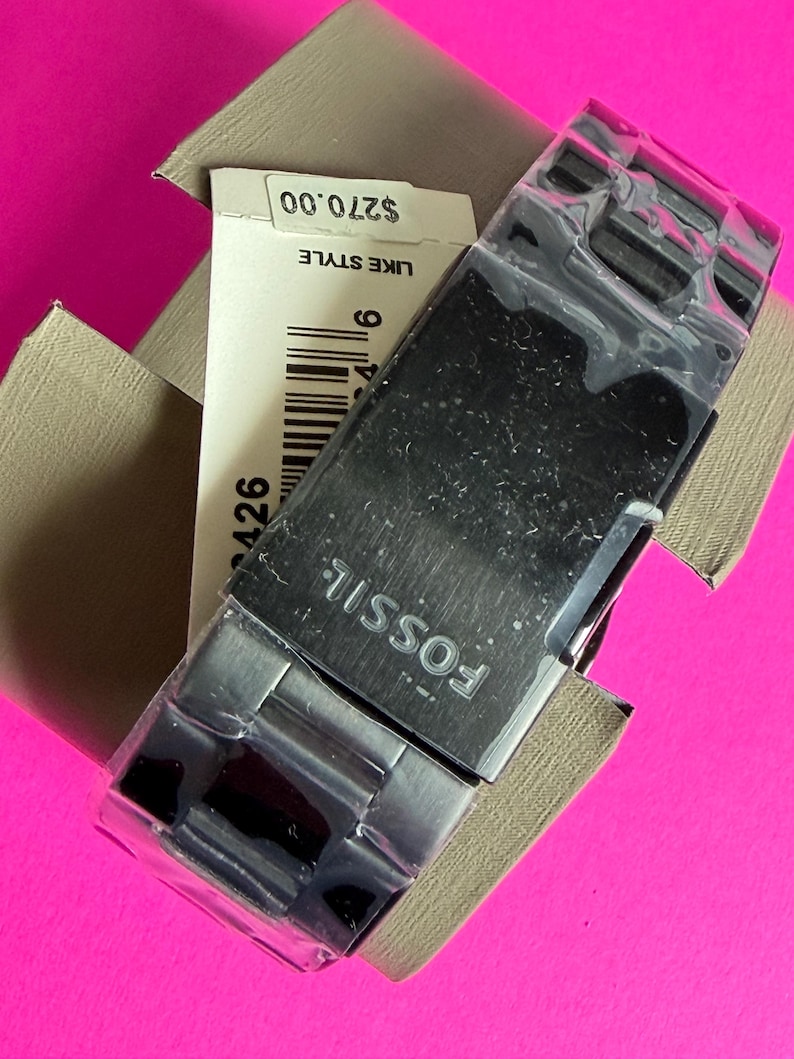 May include: A Fossil brand watch with a silver-tone metal band and a black watch face, still in its original packaging. The watch is partially covered in plastic. The watch is in a tan box with a price tag.
