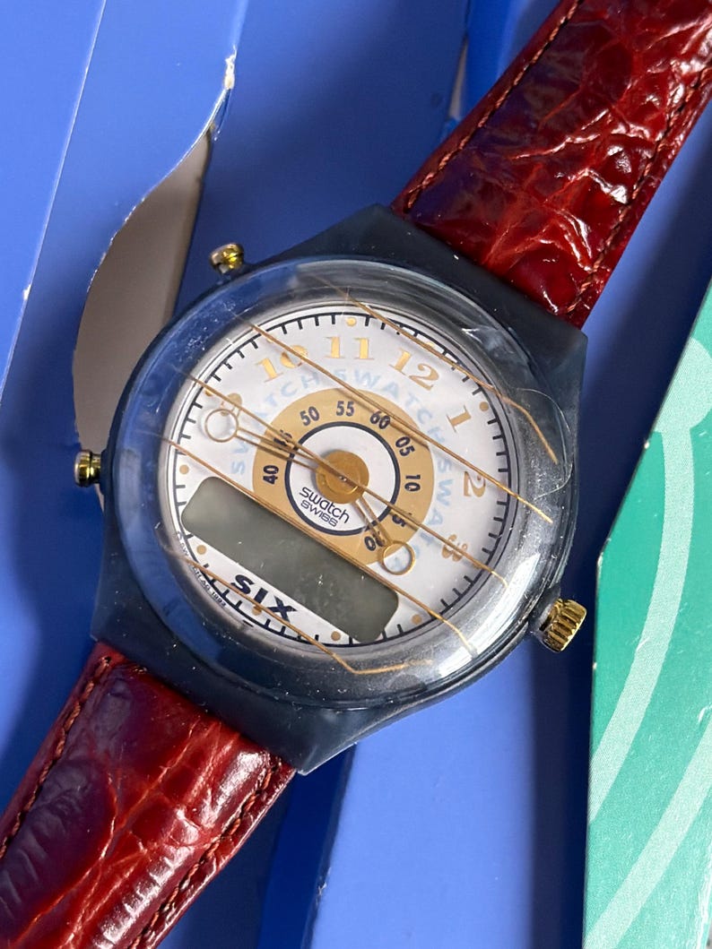 May include: A vintage Swatch watch with a white face, gold accents, and a digital display. The watch has a black case and a red leather strap. The watch face displays the text "Swatch Swiss" and "SIX".
