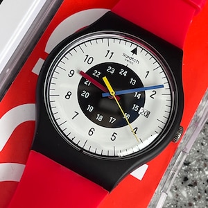 May include: A black Swatch watch with a white face, black bezel, and red strap. The watch face features black, yellow, blue, and red hands, with the Swatch Swiss logo. The watch is on a red background.