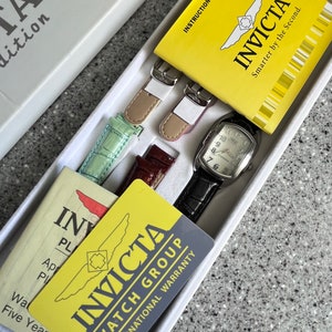 Puede incluir: Una caja de reloj Invicta amarilla y negra con un reloj plateado, dos correas de reloj de cuero marr&oacute;n y dos correas de reloj verde y rosa. La caja incluye un manual de instrucciones amarillo y una tarjeta de garant&iacute;a amarilla con el texto "INVICTA WATCH GROUP INTERNATIONAL WARRANTY" y "INVICTA Smarter by the Second."