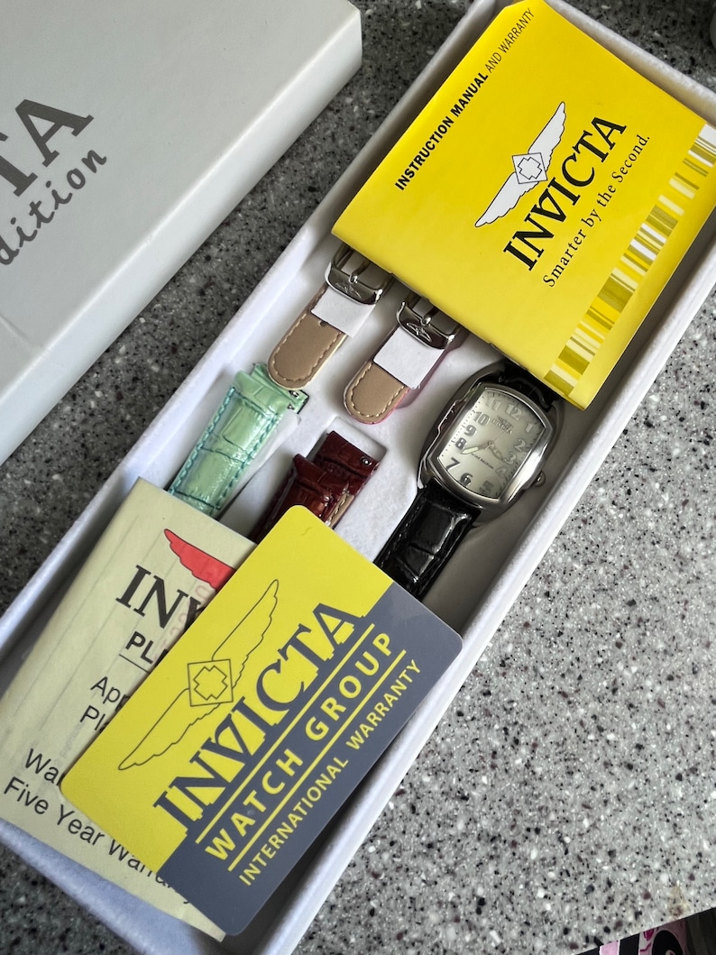 Puede incluir: Una caja de reloj Invicta amarilla y negra con un reloj plateado, dos correas de reloj de cuero marr&oacute;n y dos correas de reloj verde y rosa. La caja incluye un manual de instrucciones amarillo y una tarjeta de garant&iacute;a amarilla con el texto "INVICTA WATCH GROUP INTERNATIONAL WARRANTY" y "INVICTA Smarter by the Second."