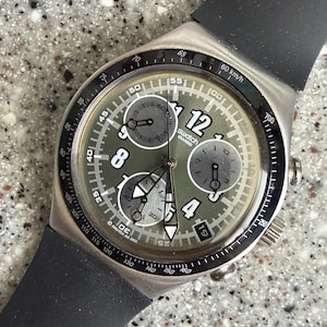May include: A Swatch chronograph watch with a matte olive green dial, black rubber strap, and silver-tone case. The dial features three sub-dials, white Arabic numerals at 4, 8, and 12, a date window, and a black tachymeter bezel marked 60 km/h.