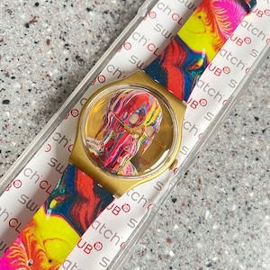 May include: A gold-toned Swatch watch with a colorful abstract design on the face and strap. The watch face features a vibrant, multi-colored image. The strap displays a swirling pattern of red, yellow, and pink.
