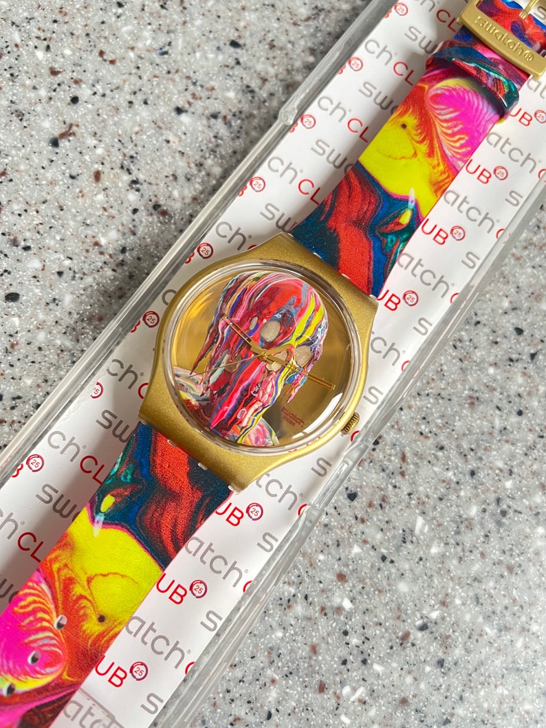 May include: A gold-toned Swatch watch with a colorful abstract design on the face and strap. The watch face features a vibrant, multi-colored image. The strap displays a swirling pattern of red, yellow, and pink.