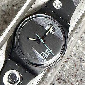 Puede incluir: Reloj Swatch negro con un diseño geométrico en la esfera y la correa. La esfera del reloj presenta un fondo negro con formas geométricas blancas y grises, y agujas azul claro. La correa tiene un patrón repetitivo de formas blancas y negras.