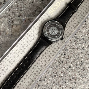 Puede incluir: Un reloj de pulsera plateado con una correa de tela negra y costuras blancas. La esfera del reloj tiene n&uacute;meros y manecillas negros, y las palabras "Stop-watch" est&aacute;n impresas en la correa.