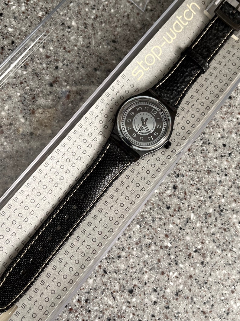Puede incluir: Un reloj de pulsera plateado con una correa de tela negra y costuras blancas. La esfera del reloj tiene n&uacute;meros y manecillas negros, y las palabras "Stop-watch" est&aacute;n impresas en la correa.