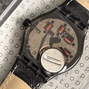 Puede incluir: Un reloj negro con una banda de tela negra y una tapa de bater&iacute;a plateada. La esfera del reloj es visible y tiene un fondo plateado con marcas negras. El reloj est&aacute; en una caja con la palabra "stop" repetida muchas veces.