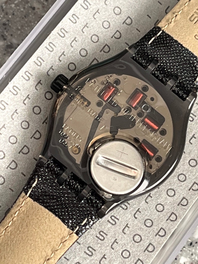Puede incluir: Un reloj negro con una banda de tela negra y una tapa de bater&iacute;a plateada. La esfera del reloj es visible y tiene un fondo plateado con marcas negras. El reloj est&aacute; en una caja con la palabra "stop" repetida muchas veces.