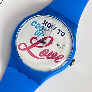 May include: A blue Swatch watch with a white face and the text "How to Cook Up Love" in pink and blue lettering. The watch has a clear plastic face and a silver second hand.