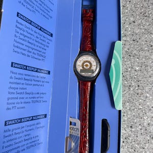 May include: A Swatch BeepUp Numeric watch in its original packaging. The watch has a red leather band and a round face with a white dial and black numerals. The packaging is blue with text in multiple languages.