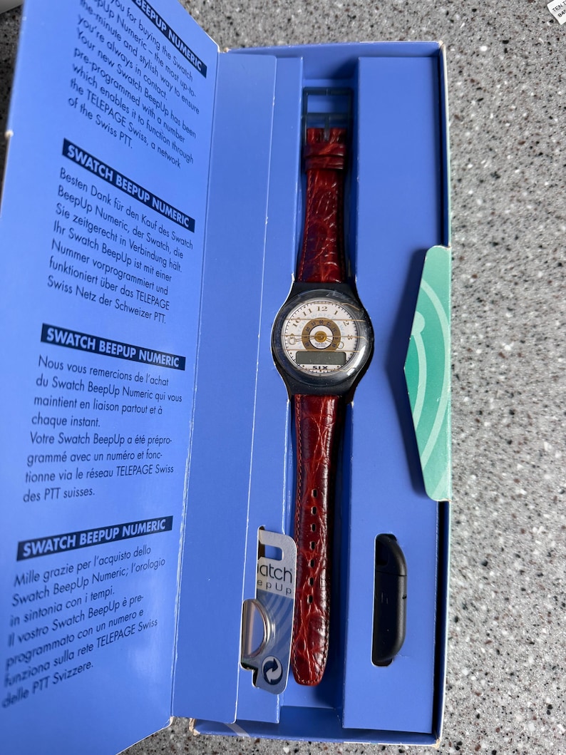 May include: A Swatch BeepUp Numeric watch in its original packaging. The watch has a red leather band and a round face with a white dial and black numerals. The packaging is blue with text in multiple languages.