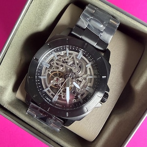 May include: A black Fossil automatic watch with a skeleton dial, showcasing the inner workings. The watch is in its original box, with a black metal band and white hands. The watch is new and has protective plastic on the band.