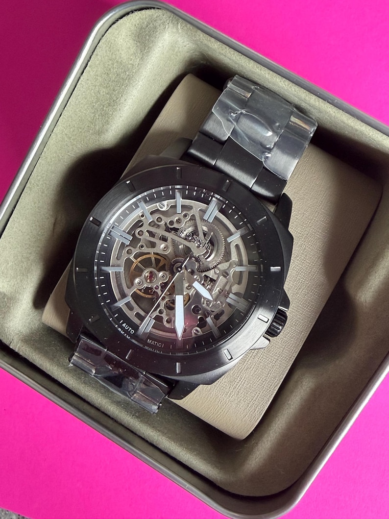 May include: A black Fossil automatic watch with a skeleton dial, showcasing the inner workings. The watch is in its original box, with a black metal band and white hands. The watch is new and has protective plastic on the band.
