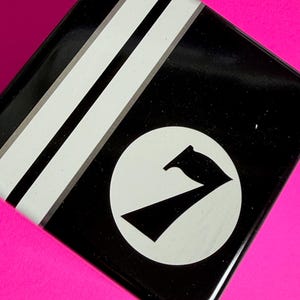 May include: Black square tin with white stripes and a white circle containing a black stylized number seven. The tin sits on a bright pink surface. The design is simple and graphic.