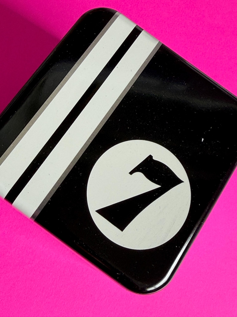 May include: Black square tin with white stripes and a white circle containing a black stylized number seven. The tin sits on a bright pink surface. The design is simple and graphic.