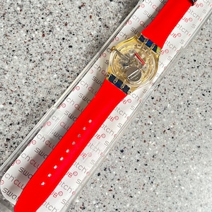 May include: A new Swatch watch with a transparent face and a bright red silicone band. The watch has a gold-colored case and a colorful floral buckle. The watch is in its original packaging.