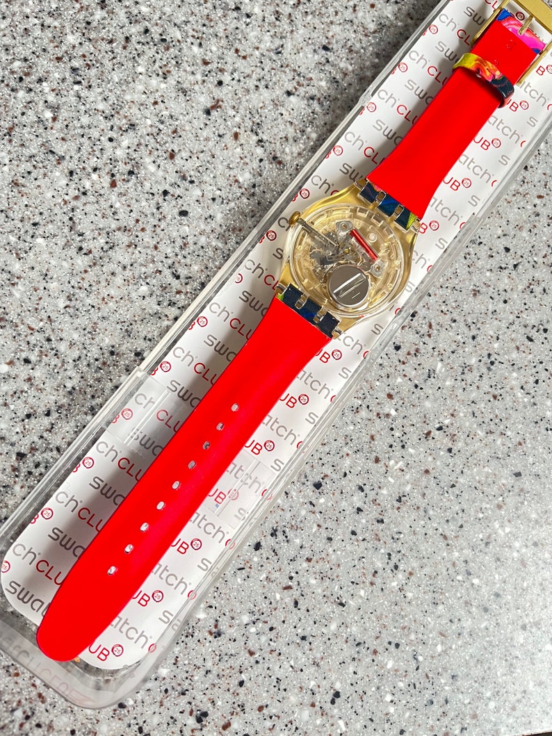May include: A new Swatch watch with a transparent face and a bright red silicone band. The watch has a gold-colored case and a colorful floral buckle. The watch is in its original packaging.