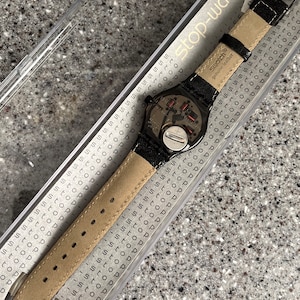 Puede incluir: Un reloj Swatch negro con una correa de cuero negro y una esfera plateada. El reloj est&aacute; en una caja de pl&aacute;stico transparente con las palabras "stop-watch" impresas en el lateral. El reloj tiene un peque&ntilde;o bot&oacute;n plateado en el lateral y las palabras "Swatch" y "cuero genuino" impresas en la correa.