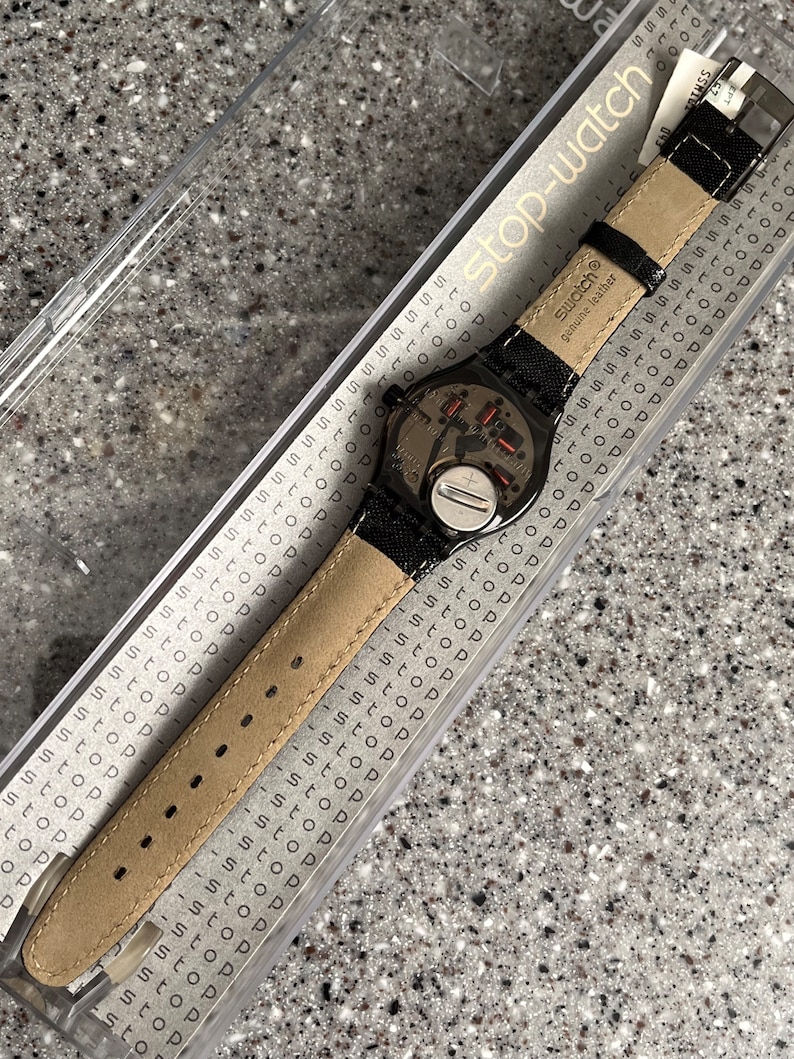 Puede incluir: Un reloj Swatch negro con una correa de cuero negro y una esfera plateada. El reloj est&aacute; en una caja de pl&aacute;stico transparente con las palabras "stop-watch" impresas en el lateral. El reloj tiene un peque&ntilde;o bot&oacute;n plateado en el lateral y las palabras "Swatch" y "cuero genuino" impresas en la correa.