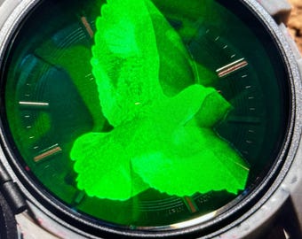 Puede incluir: Primer plano de una esfera de reloj con una silueta de pájaro verde en el centro. La esfera del reloj está rodeada por un bisel negro y una correa blanca.