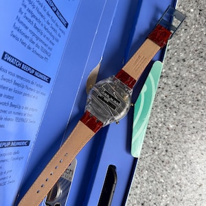 May include: A Swatch BeepUp Numeric watch with a clear plastic case and a red leather strap. The watch is displayed in its original blue box, with text in multiple languages visible on the box.