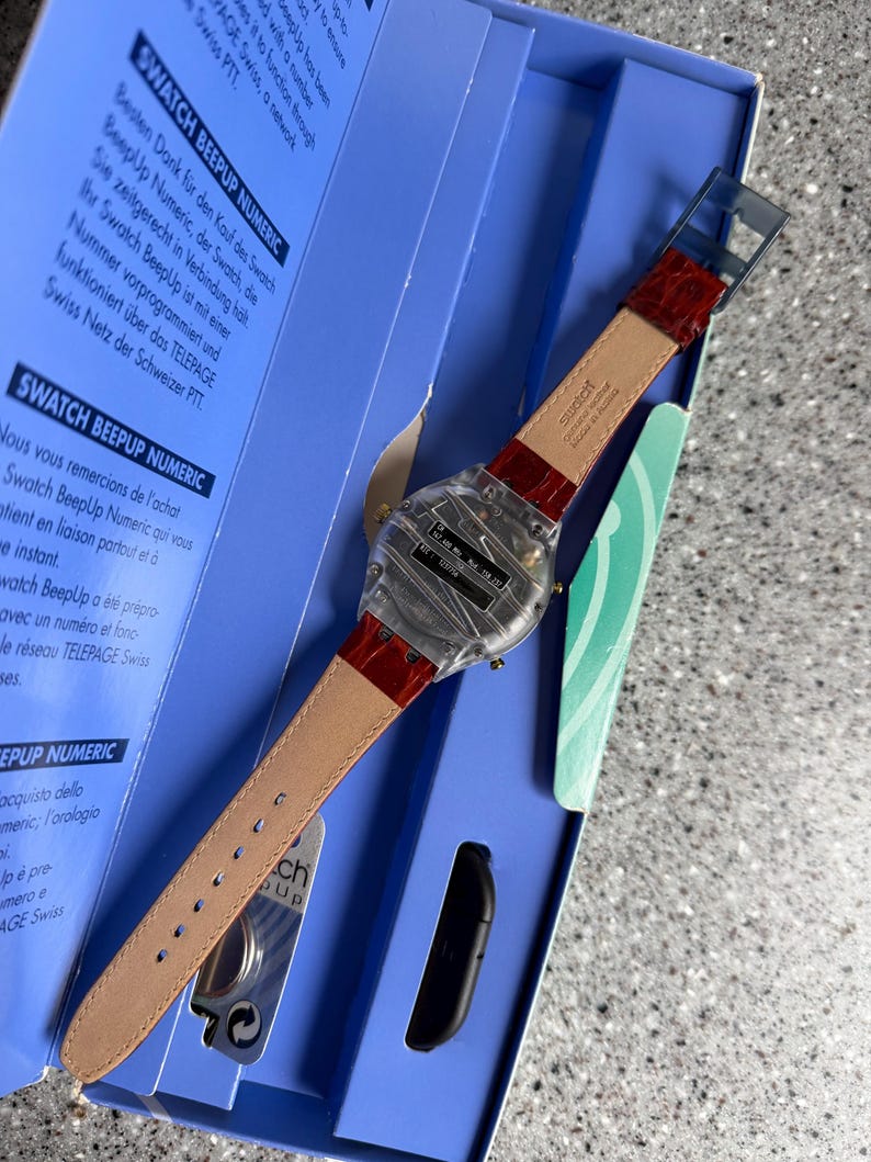 May include: A Swatch BeepUp Numeric watch with a clear plastic case and a red leather strap. The watch is displayed in its original blue box, with text in multiple languages visible on the box.