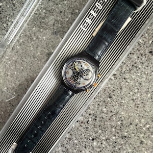 May include: A black and silver Swatch watch with a blue leather band. The watch face features a world map and the text "Chrono Neo" is printed on the band.