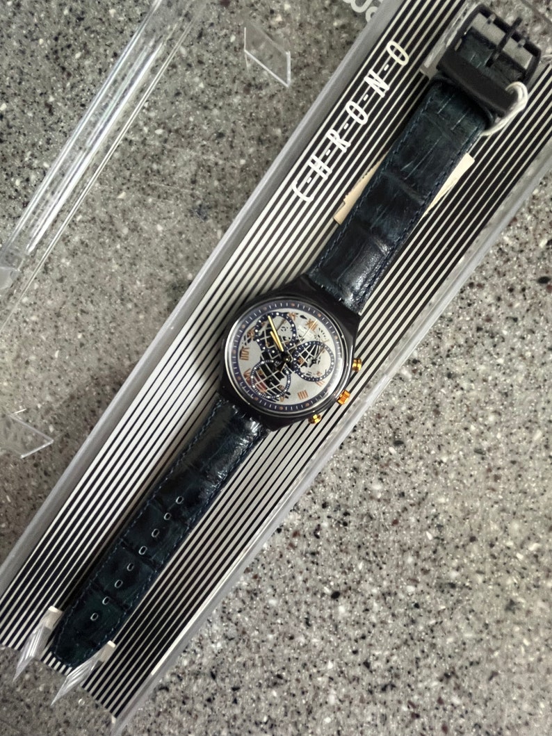 May include: A black and silver Swatch watch with a blue leather band. The watch face features a world map and the text "Chrono Neo" is printed on the band.