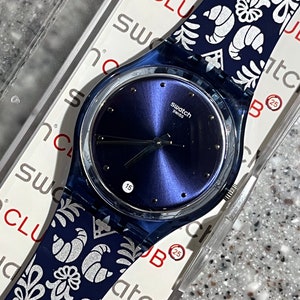May include: A blue Swatch watch with a dark blue face and a patterned strap featuring white floral and croissant designs. The watch face displays the date 15. The watch is in its original packaging.