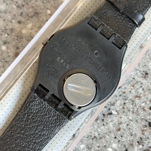 K&ouml;nnte beinhalten: Schwarze Armbanduhr mit strukturiertem Lederarmband. Das Zifferblatt ist schwarz mit den Worten "PATENTED WATERRESISTANT" und "SWISS MADE" aufgedruckt. Die Uhr hat ein silbernes Batteriefach.