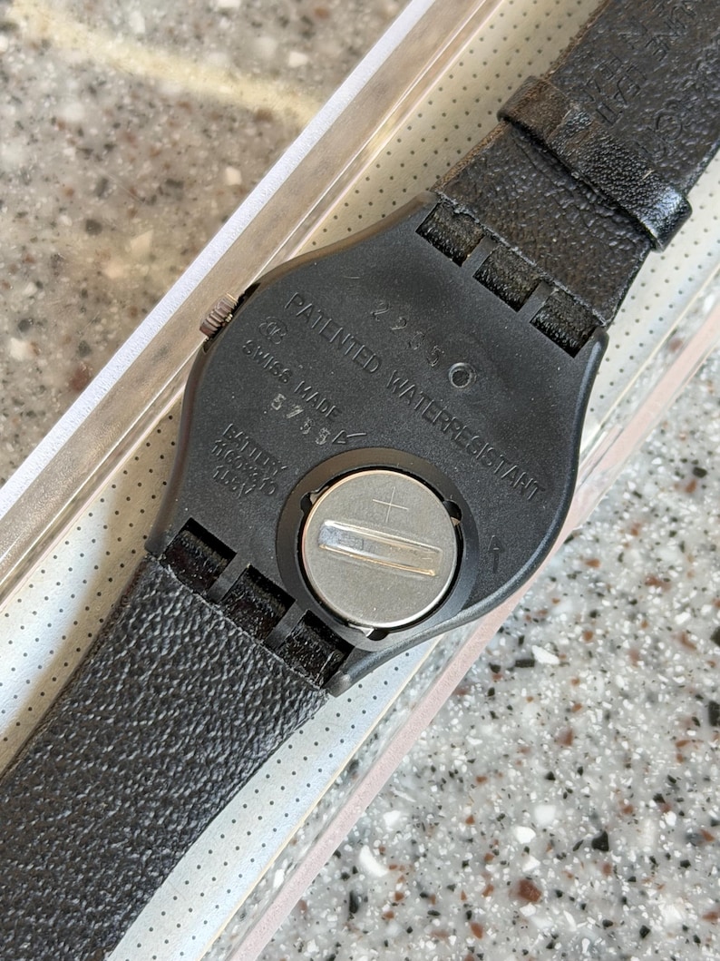 K&ouml;nnte beinhalten: Schwarze Armbanduhr mit strukturiertem Lederarmband. Das Zifferblatt ist schwarz mit den Worten "PATENTED WATERRESISTANT" und "SWISS MADE" aufgedruckt. Die Uhr hat ein silbernes Batteriefach.