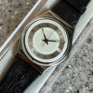 Może przedstawiać: Zegarek Swatch ze srebrną kopertą i czarnym skórzanym paskiem. Tarcza zegarka jest biała z czarnymi rzymskimi cyframi i wskazówkami. Zegarek jest w oryginalnym opakowaniu.